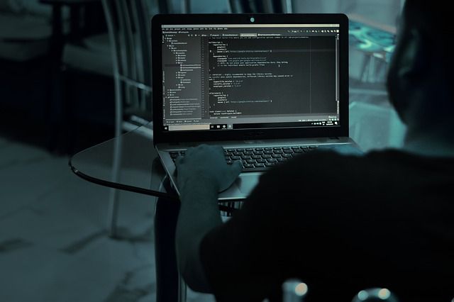 Ein Mann sitzt im dunklen Raum an einem Glastisch vor seinem Laptop und programmiert. Der Mann ist nur von hinten zu sehen, der Blick auf den Laptop ist frei. Es ist Code sehen, hier: Cyberversicherung
