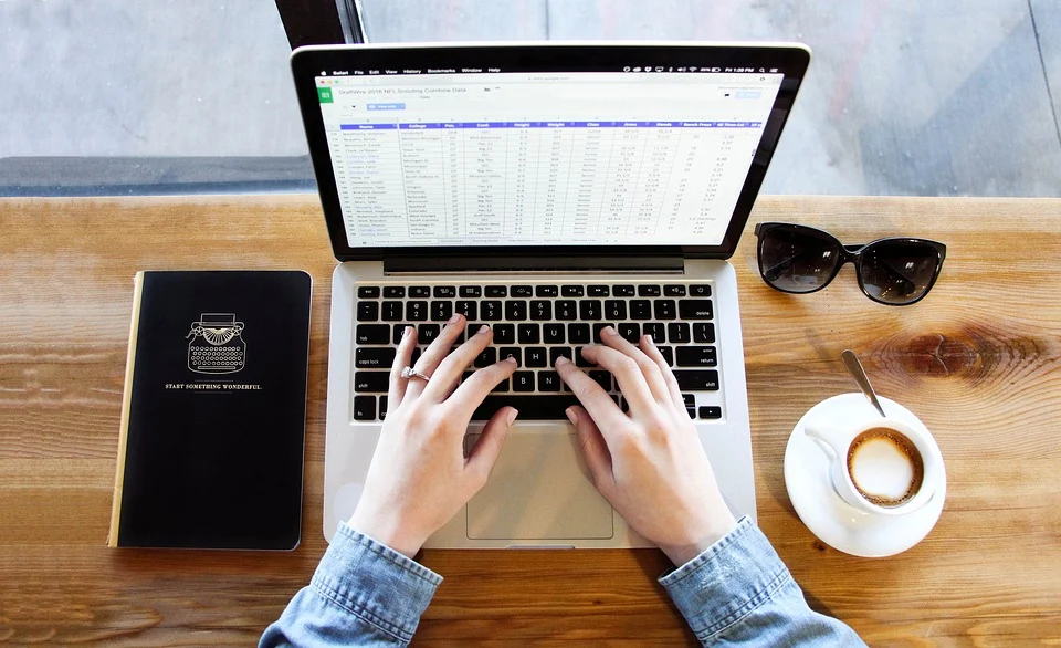Frau am Laptop beim Rechnung schreiben mit Rechnungsprogramm, daneben einen Kaffee, ein Notizbuch und eine Sonnenbrille.