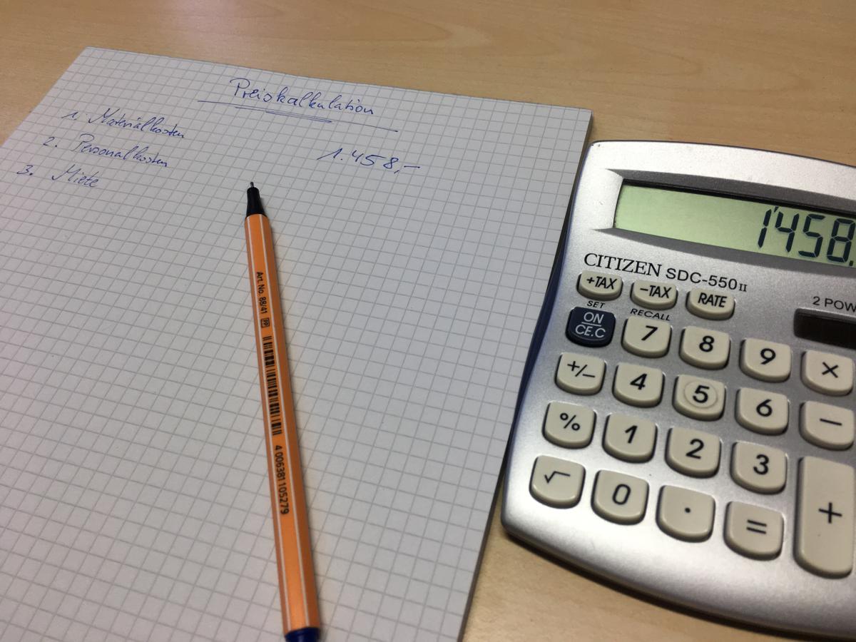 Ein blauer Faserstift liegt auf einem karierten Block Papier. Mit der Preiskalkulation wurde bereits begonnen, Materialkosten, Personalkosten. Ein Taschenrechner liegt daneben die erste Zahl eingetippt wurde.