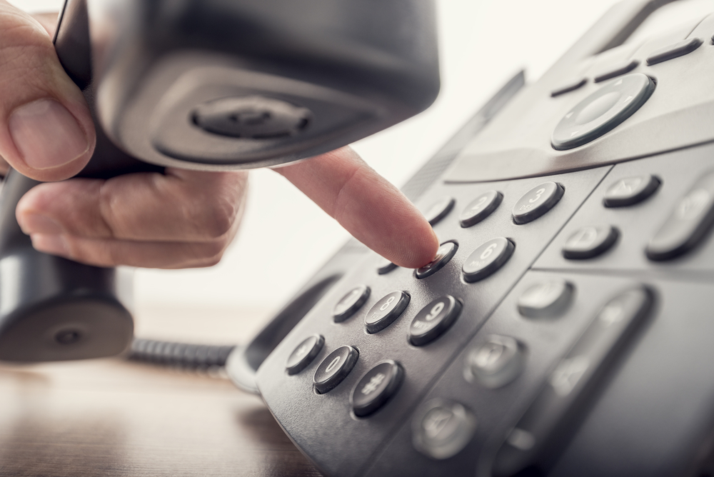 Eine Hand hält einen Telefonhörer fest und drückt mit dem Zeigefinger auf der Tastatur des Telefons eine, hier: Nachfassen am Telefonangebote zu Aufträgen machen